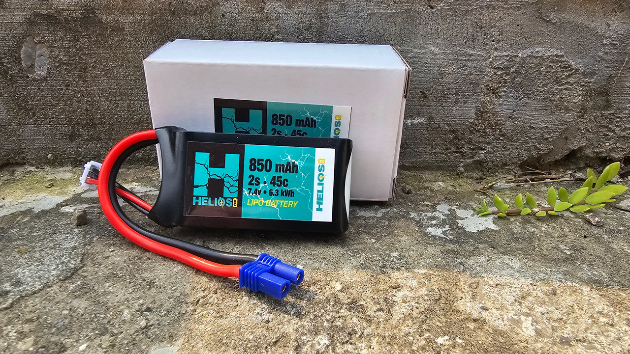 A 2S 850 mAh 45c Lipo battery with a blue label on it, resting on a stone surface with a connected wire.