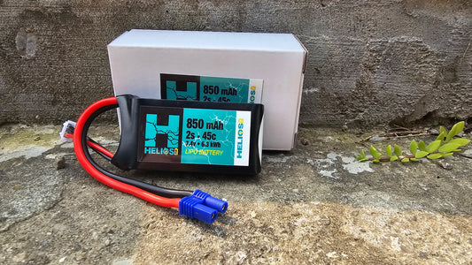 A 2S 850 mAh 45c Lipo battery with a blue label on it, resting on a stone surface with a connected wire.