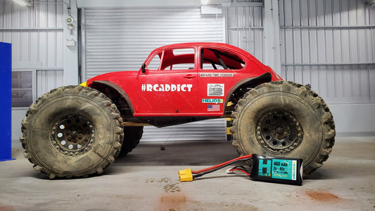 A red remote control car with 'H3Crazy' written on the side, featuring large off-road tires, parked inside a warehouse with a battery pack and charger on the floor.